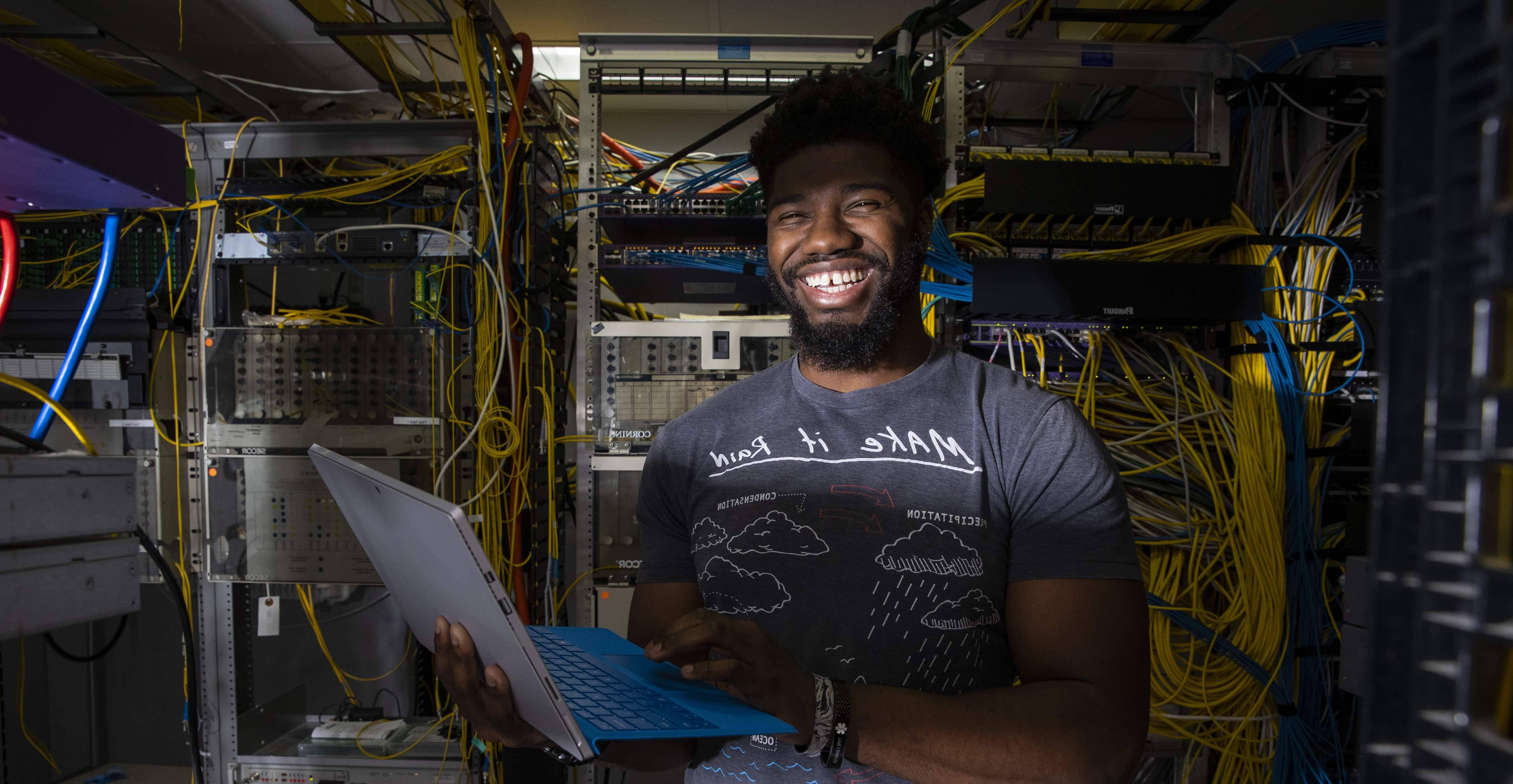 Student with laptop smiling in server room with cables all around.